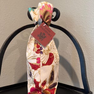 May include: A wine bottle bag with a floral pattern in red, pink, and gold on a cream background. The bag has a tag that reads "Wine Passport". The bag is tied at the top and sits on a black metal stand.