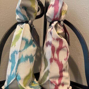 May include: Two fabric wine bottle bags with abstract patterns. One bag features blue and green brushstrokes, the other pink and purple. Each bag is tied with a black band and a decorative button. The bags are displayed on a black metal stand.