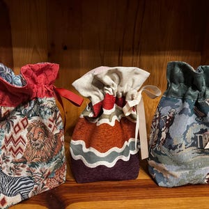 May include: Three decorative drawstring bags with unique tapestry patterns. The bags feature a red, orange, and blue color scheme. One bag has a lion and zebra design, another has a wavy pattern, and the third has a landscape scene.