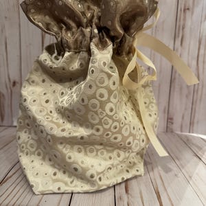 May include: A beige drawstring bag with a repeating pattern of circles. The top of the bag is a darker brown with a similar pattern. A cream-colored ribbon is tied in a bow on the side.