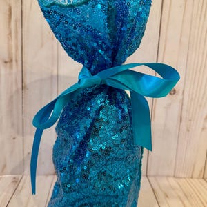 May include: A turquoise sequined gift bag tied with a matching satin ribbon. The bag is filled and gathered at the top, creating a decorative presentation. The sequins shimmer, adding a touch of elegance.
