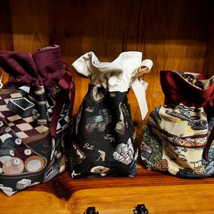 May include: Three drawstring bags with different patterned fabrics. One bag features a pool table design, another has a tractor and golf theme, and the third displays an airplane and landscape scene. Each bag has a contrasting burgundy top and ribbon.