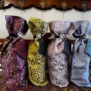 May include: Four decorative wine bottle bags in shades of purple, green, and blue. Each bag is tied with a black ribbon and a silver embellishment. Tags reading "Wine Passport" are attached.