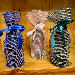 May include: Three decorative wine bottle bags. The bags are made of sequined fabric in blue, pink, and teal. Each bag is tied with a matching satin ribbon. The bags are designed to hold a standard wine bottle.