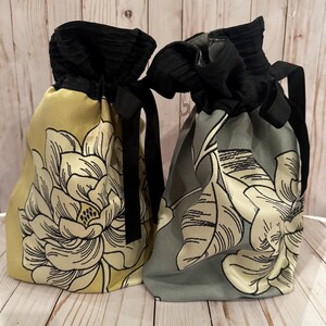 May include: Two drawstring bags with floral designs. One bag is yellow with a black and white floral pattern, and the other is gray with a similar design. Both have black velvet tops and black ribbon ties.