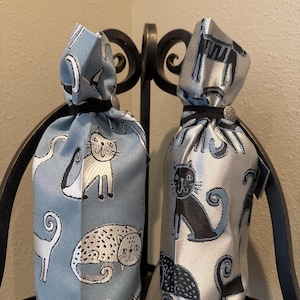 May include: Two fabric bottle bags with cat designs. One bag is light blue with white and black cat illustrations. The other is silver with black cat illustrations. Both bags are tied with black cords and decorative clasps.