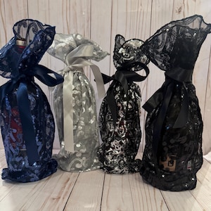 May include: Four decorative wine bottle covers. The covers are made of sequined fabric in navy blue, silver, black, and black. Each bottle is tied with a satin ribbon in a matching color. The bottles are on a light wood surface.
