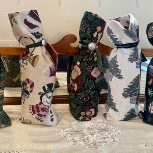 May include: Five decorative wine bottle bags with festive holiday patterns. Designs include holly, snowmen, Santa Claus, Christmas trees, and snowflakes. Each bag is tied with a ribbon and a decorative button.