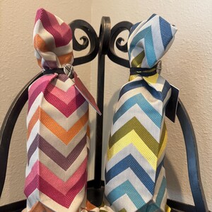 May include: Two wine bottle gift bags with chevron patterns. One bag features pink, orange, and purple chevrons, while the other has blue, green, and yellow chevrons. Each bag is tied at the top with a black elastic band and a small tag.