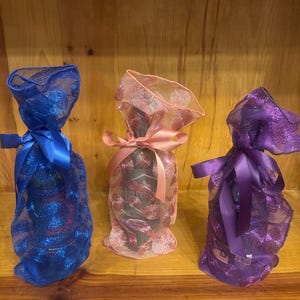 May include: Three bottles of wine are wrapped in decorative mesh bags. The bags are blue, pink, and purple, each tied with a matching satin ribbon. The bottles are on a wooden surface.