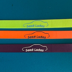 May include: Three keychains with different coloured straps: lime green, orange, and purple. Each keychain has a silver key ring and a white outline of a car with the text "Send Ludes" printed on the strap.