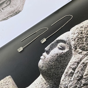 May include: A pair of silver threader earrings with clear, rectangular crystal accents. The earrings are displayed on a dark surface with a stone sculpture in the background. The earrings have a delicate chain and a straight post.