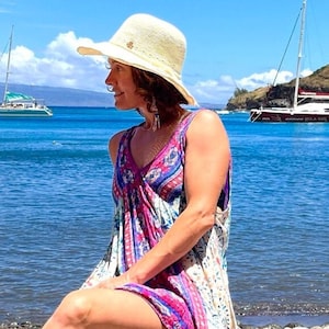 May include: A woman wearing a white sun hat and a floral dress sits on a rocky beach, looking out at the ocean. Two sailboats are anchored in the distance.