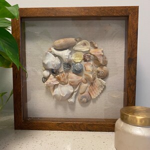May include: A framed square artwork featuring a circular arrangement of various seashells on a beige fabric background. The frame is made of dark wood with a glass front.