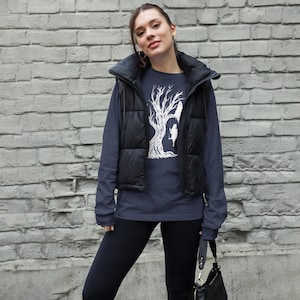 May include: A navy blue long-sleeved shirt with a white graphic of a tree with two birds perched on branches. The shirt is worn by a person with a black puffer vest and black leggings.
