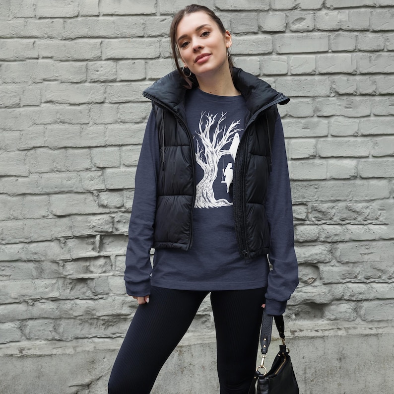 May include: A navy blue long-sleeved shirt with a white graphic of a tree with two birds perched on branches. The shirt is worn by a person with a black puffer vest and black leggings.