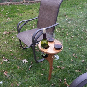 May include: A wooden wine holder with two glass holders and a bottle holder. The holder is on a single leg and is placed on the grass next to a brown patio chair.
