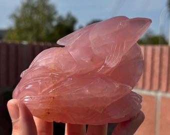 Puede incluir: Una escultura tallada de un dragón de cuarzo rosa. El dragón está enroscado con la cabeza apoyada en las patas. La escultura está hecha de cuarzo rosa translúcido con un ligero brillo.