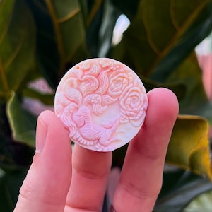 May include: A round, pink and white shell cameo with a carved floral design. The cameo is held in a hand against a green leafy background.