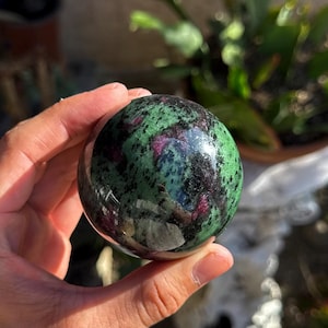 May include: A polished spherical gemstone featuring a mottled pattern of deep forest green and vibrant magenta-pink mineral inclusions. The surface is smooth and reflective, showcasing a natural contrast between the dark green and bright pink areas.