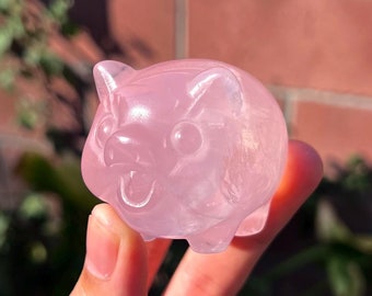 May include: A pink crystal pig figurine carved from rose quartz. The figurine is smooth and polished, with a translucent appearance.
