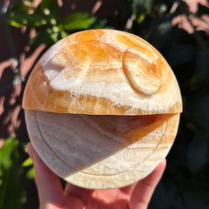 Puede incluir: Una esfera de calcita naranja y blanca tallada con un diseño de luna. La esfera está dividida en dos mitades y las dos mitades encajan.