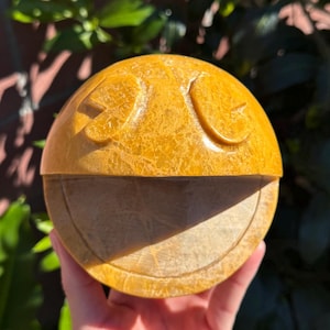 以下が含まれることがあります： 笑顔の顔をした、黄色い丸い石の彫刻。彫刻は、ざらざらした質感の明るい色の石で作られています。