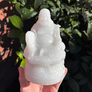 Könnte beinhalten: Eine weiße Buddha-Statue, die lächelt und in Meditation sitzt. Die Figur hat einen runden Bauch und einen ruhigen Ausdruck. Die Statue wird in einer Hand gehalten, mit einem Hintergrund aus grünem Laub.