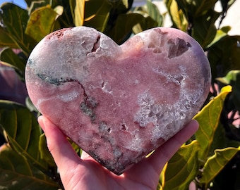 May include: A large, heart-shaped crystal in shades of pink, white, and gray is held in a hand. The crystal has a rough, textured surface with visible mineral formations. The background features green foliage and a clear blue sky.