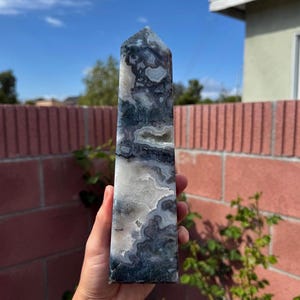 May include: A gray, white, and black banded agate obelisk crystal point. The crystal is polished and has a smooth, shiny surface.