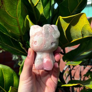 Puede incluir: Una mano sosteniendo una figurita de piedra tallada de un gato de dibujos animados de color rosa y gris. La figurita está detallada con un lazo y tiene una expresión caprichosa.