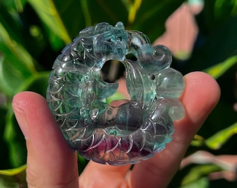 Può includere: Un ciondolo in fluorite intagliato di colore verde e blu a forma di drago. Il ciondolo è dettagliato con squame e ha un foro per un cordino.