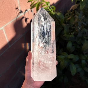 May include: A clear quartz crystal point, also known as a clear quartz wand, is a natural crystal with a pointed tip. The crystal is smooth and polished, with a translucent appearance. The crystal is held in a hand, with a brick wall and green plants in the background.