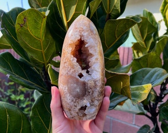 Puede incluir: Una gran geoda de color marrón claro con un centro hueco forrado de cristales blancos. La geoda se sostiene en una mano, con la abertura mirando hacia la cámara.