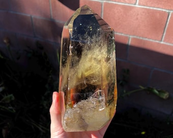 May include: A large, clear, golden citrine crystal point with a natural, raw, and rough base. The crystal is translucent and has a golden hue. The crystal is held in a hand.
