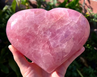 Puede incluir: Un gran cristal de cuarzo rosa en forma de corazón. El cristal es de color rosa pálido y tiene una superficie lisa y pulida.