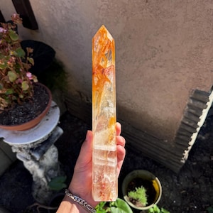 May include: A clear quartz crystal point with orange and yellow inclusions. The crystal is held up in front of a neutral background. The crystal is approximately 6 inches tall and has a pointed tip. The crystal is a natural, raw stone.