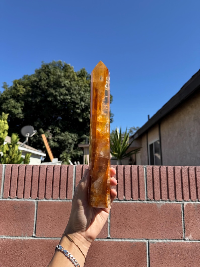 May include: A tall, amber-colored crystal point held against a blue sky. The crystal is rectangular with a pointed top and has a translucent quality. The background includes a brick wall, trees, and a house.