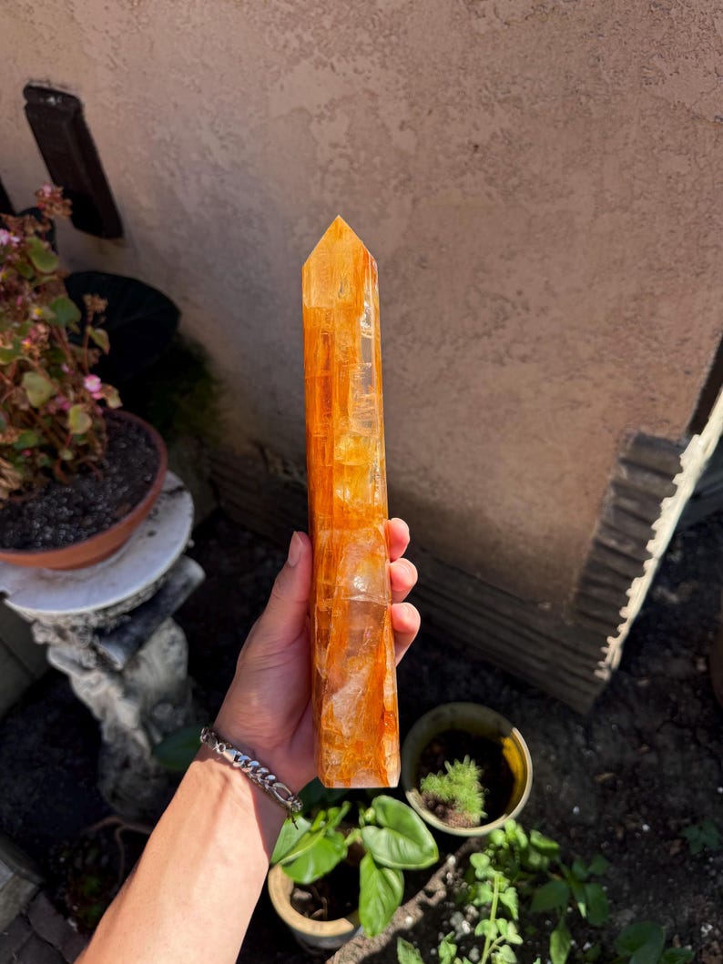 May include: A tall, orange citrine crystal point. The crystal is held in a hand and is positioned in front of a neutral background. The crystal is a vibrant, translucent orange color and has a pointed top. The crystal is a natural, raw gemstone.