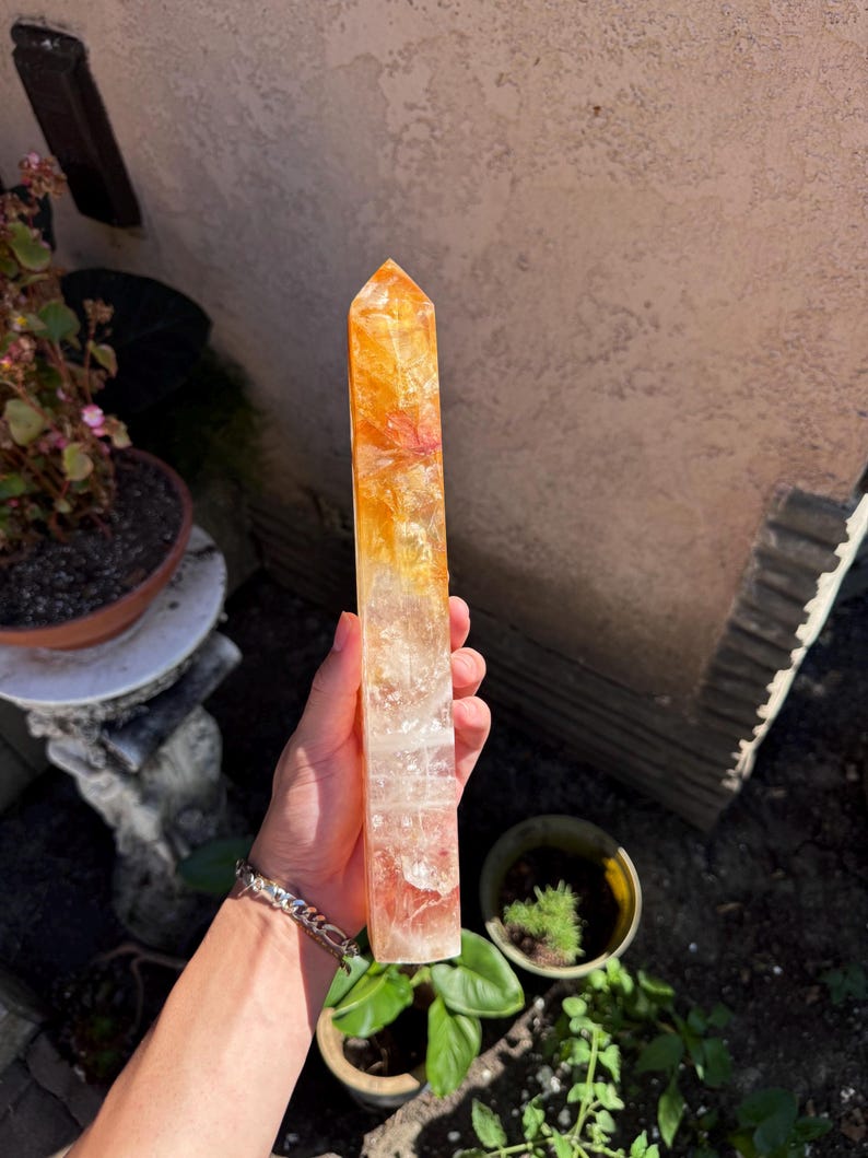 May include: A tall, rectangular crystal point with a gradient of colors, transitioning from a clear base to orange and yellow at the top. The crystal is held outdoors, with potted plants and a wall in the background. The crystal is approximately 8 inches tall.