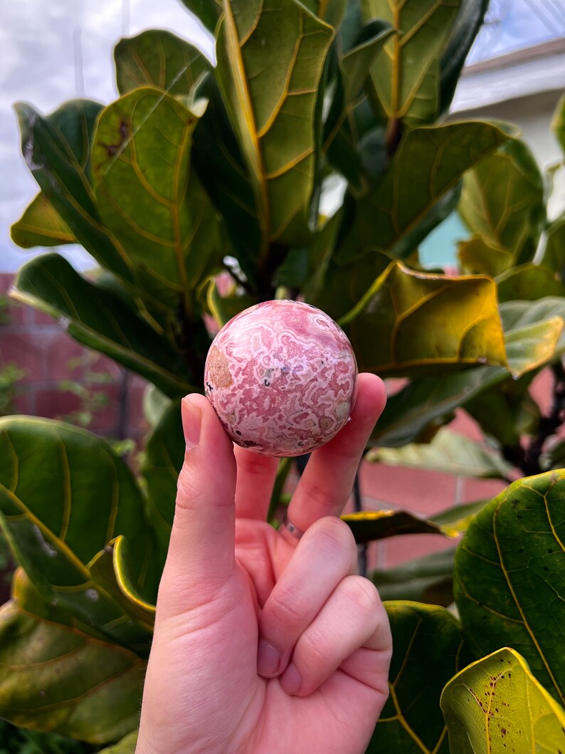 May include: A pink and white patterned sphere made of polished stone. The sphere is about the size of a golf ball.