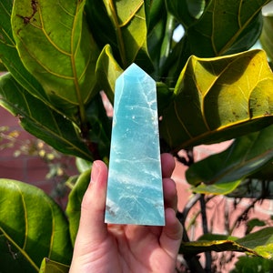 May include: A light blue aquamarine crystal point, also known as a tower or obelisk, is held in a hand. The crystal is smooth and polished, with a pointed tip and a flat base.