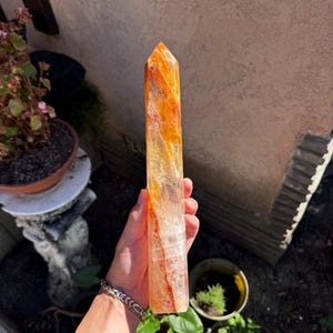 May include: A tall, orange and clear crystal point. The crystal is held in a hand and has a pointed top. The crystal has a mix of orange and clear colors. The crystal is a natural stone.