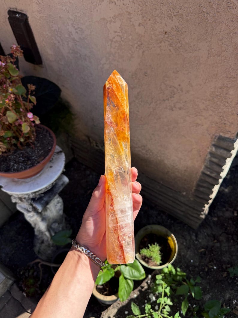 May include: A tall, orange and clear crystal point. The crystal is held in a hand and has a pointed top. The crystal has a mix of orange and clear colors. The crystal is a natural stone.