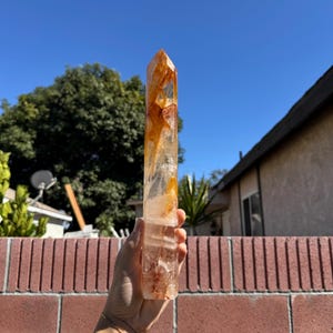May include: A clear quartz crystal point with orange and reddish-brown inclusions. The crystal is held up against a bright blue sky, showcasing its natural beauty. The crystal is a natural, unpolished gemstone.