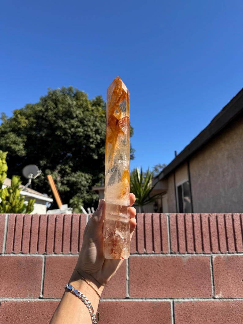 May include: A clear quartz crystal point with orange and reddish-brown inclusions. The crystal is held up against a bright blue sky, showcasing its natural beauty. The crystal is a natural, unpolished gemstone.