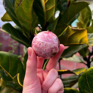 May include: A pink and white sphere with a swirling pattern, made of polished stone. The sphere is being held in a hand.