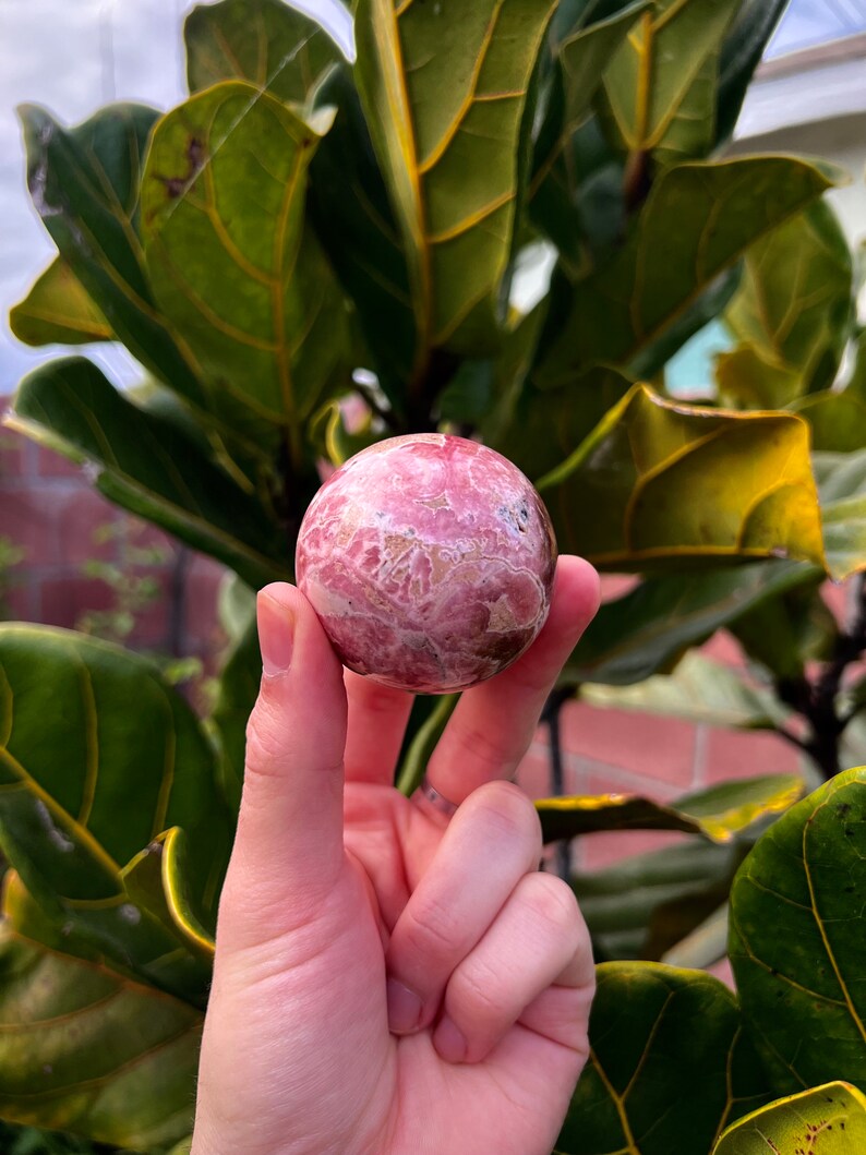 May include: A pink and white sphere with a swirling pattern, made of polished stone. The sphere is being held in a hand.