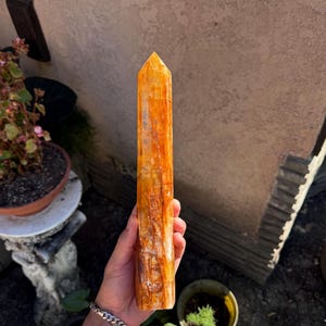 May include: A tall, orange-yellow crystal point with a polished surface. The crystal is held in a hand, showcasing its vertical lines and natural variations. The crystal's color ranges from light yellow to deep orange.