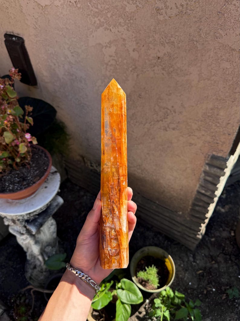 May include: A tall, orange-yellow crystal point with a polished surface. The crystal is held in a hand, showcasing its vertical lines and natural variations. The crystal's color ranges from light yellow to deep orange.